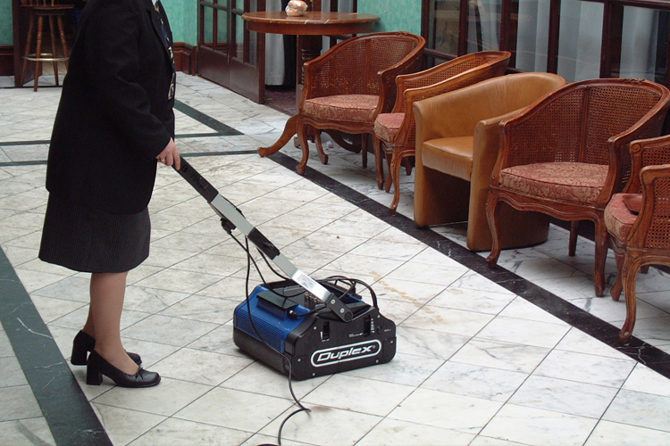 Duplex floor cleaning machine operating on a polished tiled hard floor in a commercial lobby area.