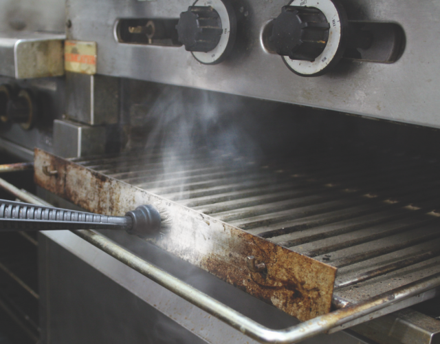 Steam cleaning an oven grill in a retirement village kitchen to remove grease and sanitise without chemicals.