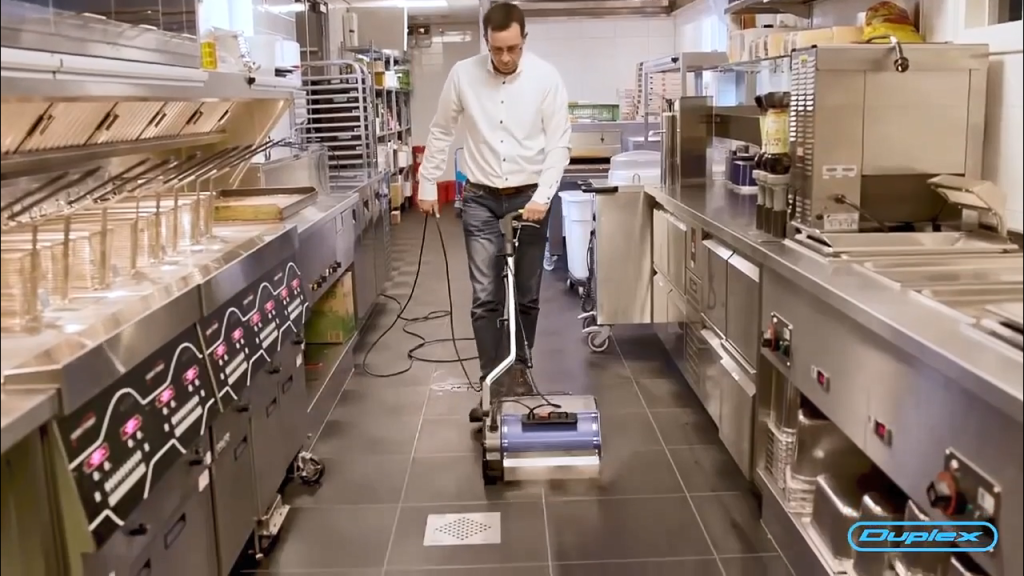 Video still showing a Duplex machine cleaning commercial kitchen tile floors between stainless counters.